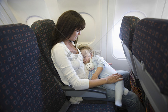 母亲和蹒跚学步的孩子在飞机上 窗户的百叶窗已经放下 蹒跚学步的孩子抱着一只玩具兔子睡着了 而妈妈则看着他们男生孩子们女性男孩们父图片