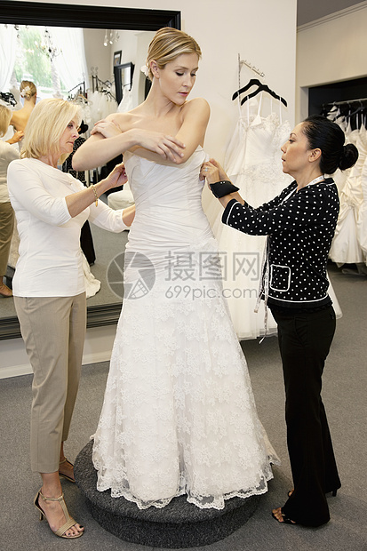 母亲和商店雇员协助年轻妇女在新娘店里穿衣服;她们帮助青年妇女图片