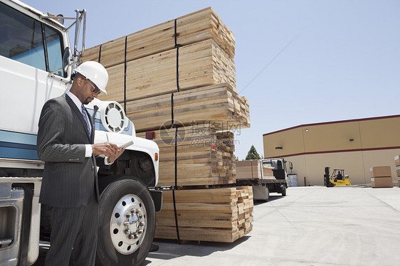 非洲裔美国男性承包商在站在伐木卡车上时使用平板电脑(Papt PC)图片