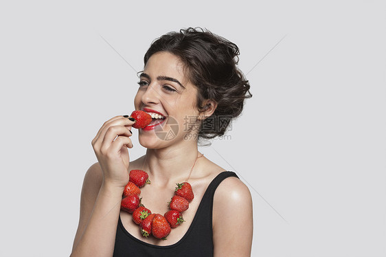 快乐的年轻女子穿着草莓项链 在灰色背景上吃一块东西时图片
