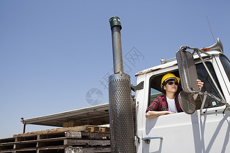 女工业工人在坐在伐木卡车上时调整镜镜的身影商业成人木材场摩托车司机女性安全帽车门陆地运输图片