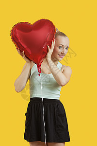 美丽的年轻女子的肖像 在黄色背景上窥视着心形气球图片