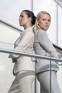 严肃的年轻女商务人士在办公室栏杆上背靠背站着女性同事玻璃阳台建筑学西装办公楼两个人生意人建筑图片