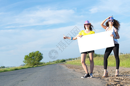 两名年轻女性站在空白的横幅上站着航程便车旅行远足者路线木板女士旅游季节女孩图片