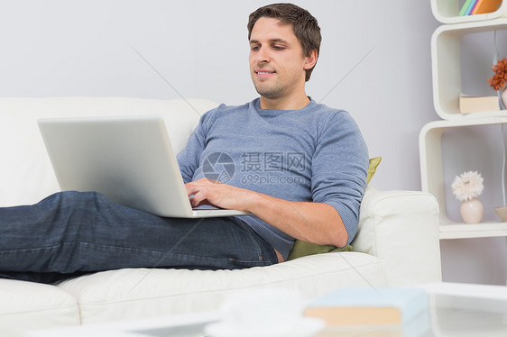 在客厅使用笔记本电脑的放松的青年男子专注长椅头发男性男人技术房子棕色牛仔裤闲暇图片