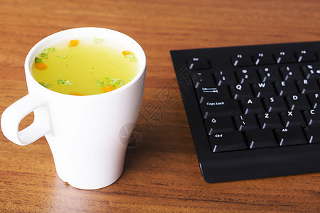 在键盘旁边的白色杯子里放热蔬菜汤图片