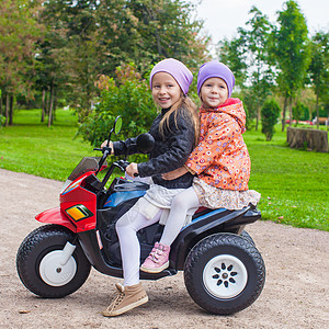 两姐妹坐在绿色公园的玩具摩托车上 坐在绿公园里晴天速度引擎裙子夹克婴儿活动微笑女孩孩子图片