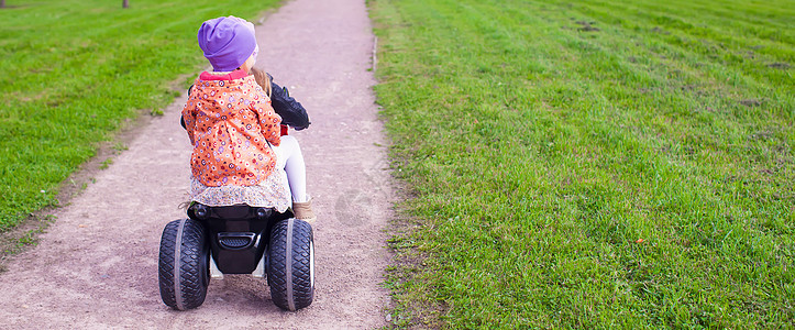 在绿公园骑摩托车的 可爱小女孩背面景色裙子孩子游戏喜悦运动引擎童年夹克姐姐晴天图片