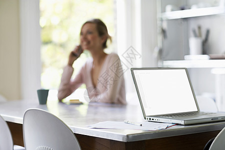 手机上的妇女坐在厨房桌上 用笔记本电脑看笔记本电脑图片