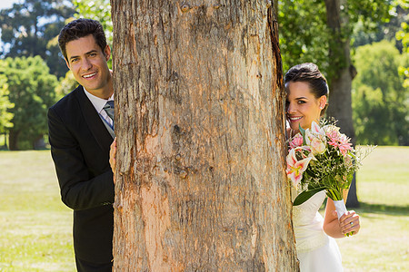 在公园树干后面的一对新婚夫妇图片