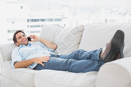 快乐的男人躺在沙发上 在电话里聊天牛仔布手机牛仔裤呼唤微笑客厅闲暇男性公寓房子图片