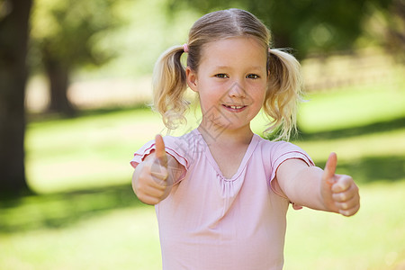 微笑的女孩在公园里挥舞着拇指绿色金发辫子绿地金发女郎闲暇童年手势浅色双手图片
