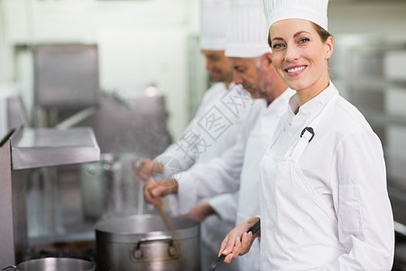 快乐的厨师在炉灶做饭时对着照相机微笑滚刀职员厨艺酒店烹饪女士炉顶团队制服合作图片