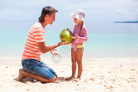 可爱小女孩和年轻父亲的肖像 在异国海滩上加椰子牛奶图片