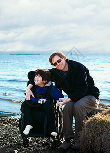 父亲与残疾儿子一起坐在湖岸边家庭孩子快乐晴天男人阳光男生轮椅需求混血儿图片