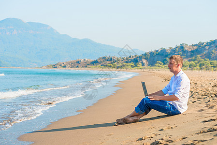 坐在沙滩上有笔记本电脑的英俊年轻男子图片