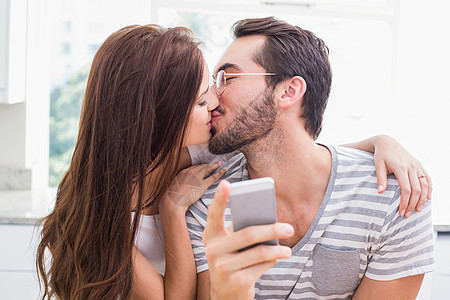 年轻男人使用智能手机 而女友亲吻他夫妻桌子女士家庭生活电话女朋友屏幕男性女性厨房图片