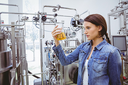 时髦的黑褐色 穿着牛仔夹克 看着啤酒杯机器职业外套生产女性工厂啤酒食品酒精增值税背景图片