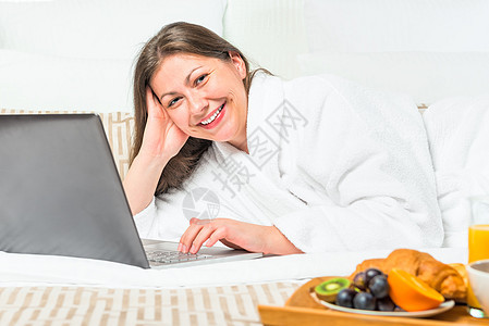 带着笔记本电脑和早餐托盘躺在床上的微笑着女人图片
