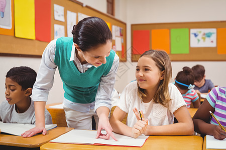 在课堂上帮助一个小女孩的老师记事本同学们混血工作笔记本快乐桌子小学生椅子写作图片