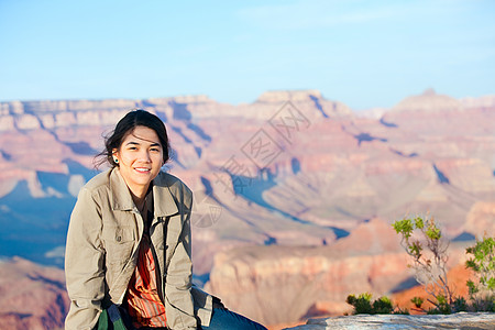 年轻女孩坐在大峡谷岩边的岩石边缘微笑晴天混血儿牛仔布女性牛仔裤石头峡谷悬崖图片