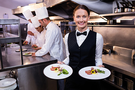 服务员向摄影机展示盘子食物领结工作烹饪女性微笑午餐厨房合作马甲图片