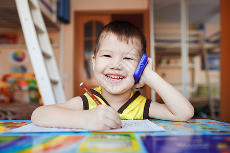 快乐的小男孩在家里用智能手机说话 同时写信图片