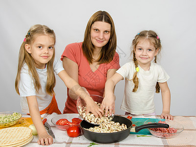 两个小女儿坐在厨房桌上 帮她妈妈把切碎的蘑菇从盘子倒到锅里图片