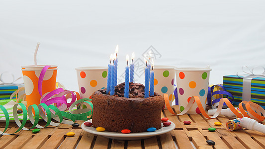 巧克力生日蛋糕 蓝色蜡烛在质朴的木桌上燃烧 背景是五颜六色的彩带 礼物 塑料杯和糖果 背景是白墙桌子日子幸福孩子们飘带盒子派对纪图片