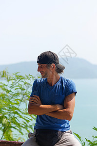 穿着白蓝T恤和黑帽子 在市公园的石栏杆上坐着 一个快乐的年轻胡子男子运动胡须男性城市男人海滩相机眼睛蓝色太阳背景图片