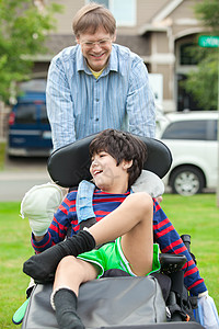父亲将10岁残疾的儿子推到户外轮椅上帮助爸爸照顾者医疗机动性男生脑瘫白色少数民族微笑图片