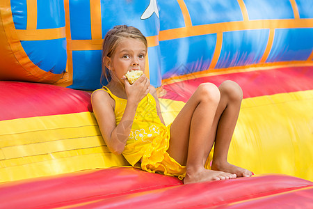 女孩坐在一个充气的蹦床入口 吃着苹果假期喘息休息公园情感娱乐童年咀嚼消遣水果图片