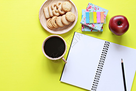 办公桌的顶部视图与空白笔记本与铅笔饼干苹果咖啡杯和黄色背景上的彩色记事本图片