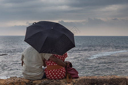 印度夫妇在海边坐着的后视线 带着雨伞看着地平线图片