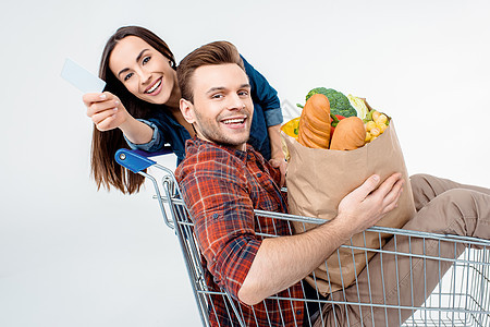 青年男子坐在装着杂货袋的购物车厢中 妇女持空白卡购物袋信用卡购物车夫妻蔬菜纸袋食物成人面包杂货图片