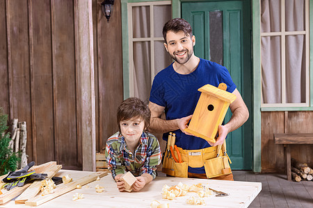 快乐的父亲和儿子拿着木鸟屋 对着镜头微笑手工爸爸领班木工作坊鸟巢男生成人男人巢箱图片