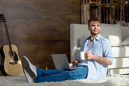 帅气的年轻人 手持笔记本电脑和杯子坐在地毯上 看远了图片