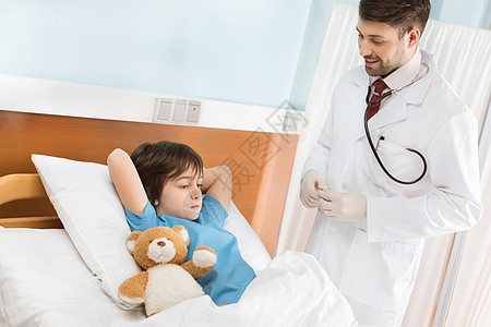 微笑儿科医生看着小男孩躺在医院床上的泰迪熊中图片