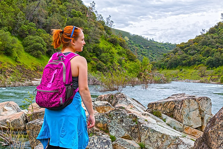女孩站在近快河岩石上闲暇天气冒险快乐山脉蓝色背包急流头发森林图片