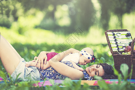年轻美丽的母亲坐在公园的毛毯上与女儿坐在一起快乐孩子篮子乐趣父母食物毯子女士说谎幸福图片