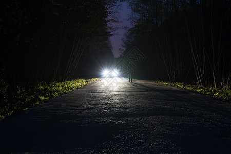 夜间穿过森林的黑暗山路边缘的男性剪影 站在路上的人对着汽车前灯 路边的车 神秘的概念 软焦点 过滤器图片