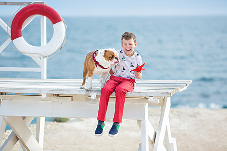 英俊的男孩少年快乐地与他的朋友斗牛犬在海边共度时光 小狗抱着玩两颗海星靠近救生圈漂浮穿着红色裤子拖鞋和 T 恤海岸宠物生活游泳孩图片