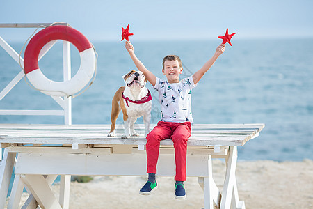 抱着海星男孩英俊的男孩少年快乐地与他的朋友斗牛犬在海边共度时光 小狗抱着玩两颗海星靠近救生圈漂浮穿着红色裤子拖鞋和 T 恤家庭游戏男生飞溅动背景