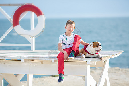 英俊的男孩少年快乐地与他的朋友斗牛犬在海边共度时光 小狗抱着玩两颗海星靠近救生圈漂浮穿着红色裤子拖鞋和 T 恤乐趣海岸磁盘椅子跑图片