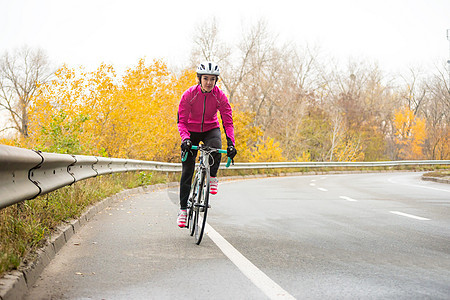 粉红夹克骑马的年轻女子 在冷秋日高速公路上的双轮赛 健康生活方式微笑速度女性头盔成人自由爱好城市活动手套图片