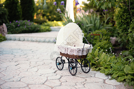 娃娃的婴儿车 古董娃娃婴儿车放置在石头人行道上 小巷在一个美丽的花园里 周围有花草树木 藤条和白色蕾丝制成的复古手推车娃娃图片