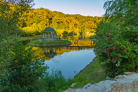 休闲凉亭位于湖心绿岛上 有桥通往 游客们欣赏夕阳照耀对岸高大树木的美景图片