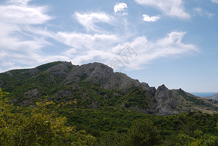 岩石山峰在一片明亮的天空背景下 在一块稀有和稠密的绿林下面的斜坡上飘浮着透明的云层图片