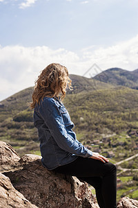 一个远足女孩坐在悬崖边缘 望着山谷和山峰的景象旅游岩石高山活动牛仔裤远足者女士石头探索游客图片