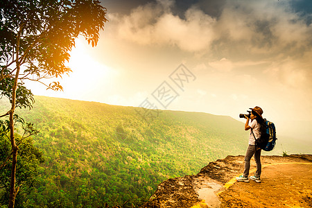 站在山崖上人背着背包 帽子和相机的年轻旅行女人站在山崖上 女摄影师在度假时拍摄热带森林 天空和云彩 亚洲女人独自旅行背景